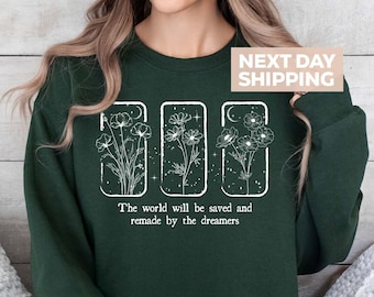 May include: Dark green sweatshirt featuring a white floral design within three rectangular frames. The text "The world will be saved and remade by the dreamers" is printed below the design. The sweatshirt is a comfortable, casual style.