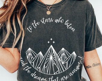 May include: A dark gray t-shirt with a white graphic design of mountains and stars. The text "to the stars who listen and the dreams that are answered" is printed on the shirt.