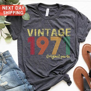 May include: A dark grey t-shirt with the text "VINTAGE 1971 Original parts" in a retro style. The shirt is displayed with blue jeans and brown sandals. The design on the shirt includes the year 1971 in red, orange, yellow, and green.