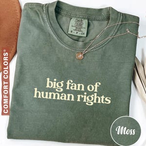 Pode incluir: Uma camiseta Comfort Colors verde musgo com o texto "big fan of human rights" em bege claro. Um colar dourado com um pequeno pingente repousa sobre a camiseta. Um chapéu marrom é visível no fundo.