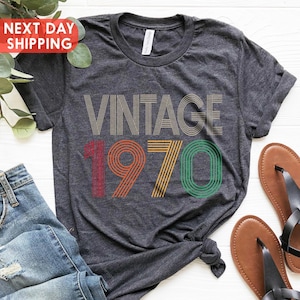 May include: Dark gray t-shirt with the text "VINTAGE 1970" in a retro font. The numbers are in red, yellow, and green. The shirt is paired with blue jeans and brown sandals with black straps. A sprig of greenery is in the background.