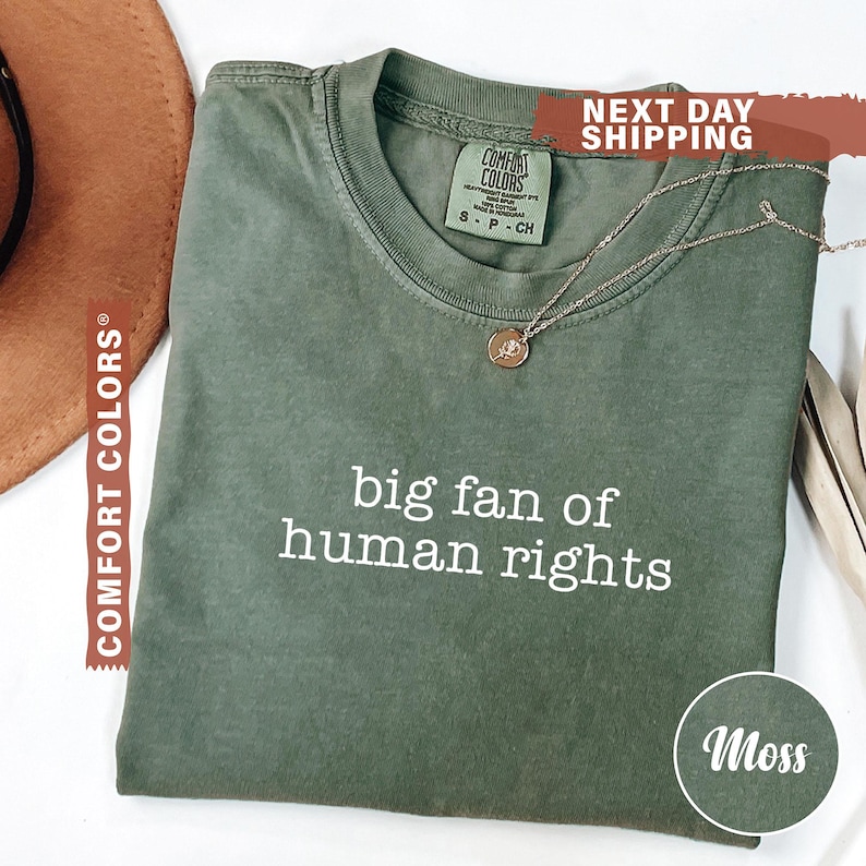 May include: A moss green t-shirt with the text "big fan of human rights" in white. The shirt has a crew neck and short sleeves. A gold necklace with a pendant is draped across the shirt. The shirt is labeled "Comfort Colors".