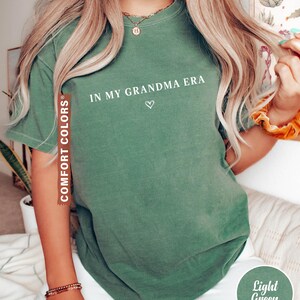 May include: Light green t-shirt with the text "IN MY GRANDMA ERA" and a small heart. The shirt is a Comfort Colors brand. The shirt is light green.