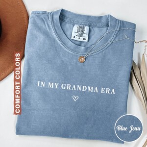 May include: Blue jean colored t-shirt with the text "IN MY GRANDMA ERA" and a heart symbol. A silver necklace with a pendant is on the shirt. The shirt is from Comfort Colors.
