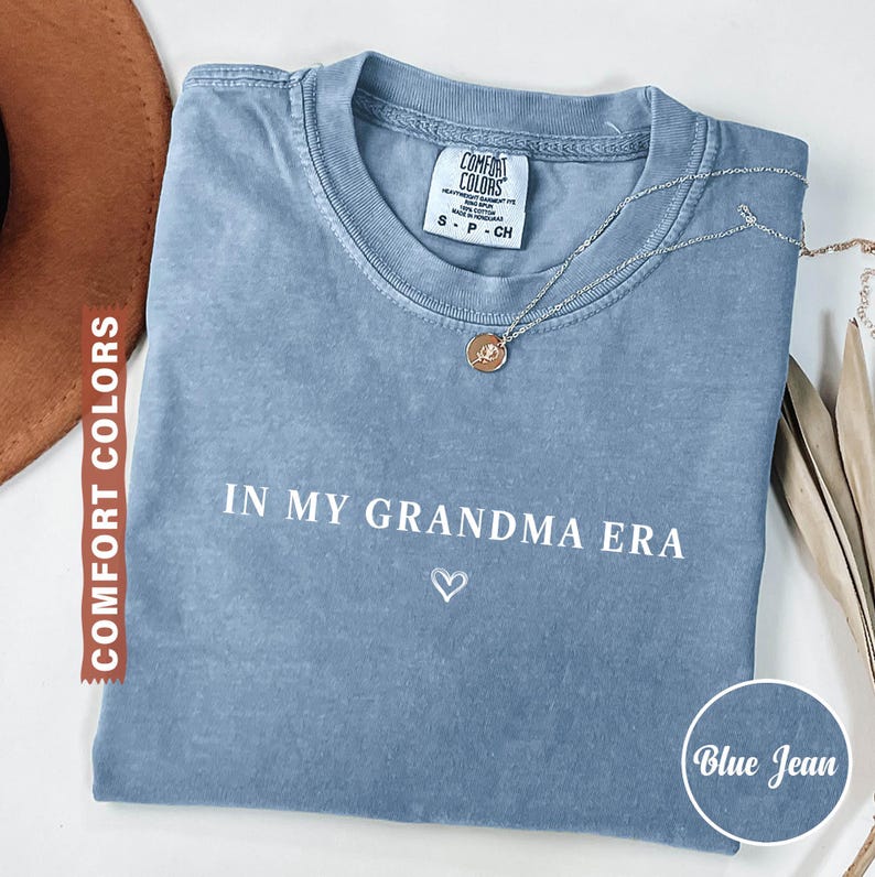 May include: Blue jean colored t-shirt with the text "IN MY GRANDMA ERA" and a heart symbol. A silver necklace with a pendant is on the shirt. The shirt is from Comfort Colors.