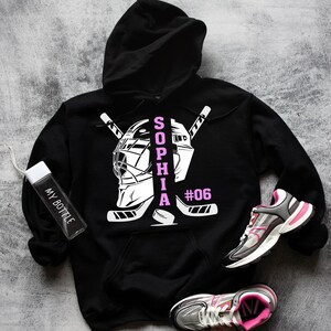 May include: Black hoodie with a white hockey goalie mask and crossed hockey sticks. The name "SOPHIA" is written vertically in pink letters and the number "#06" is written in white.