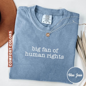 May include: A blue jean Comfort Colors t-shirt with the text "big fan of human rights" in white. A silver necklace with a pendant is on the shirt. The shirt is folded and displayed with a brown hat and dried leaves.