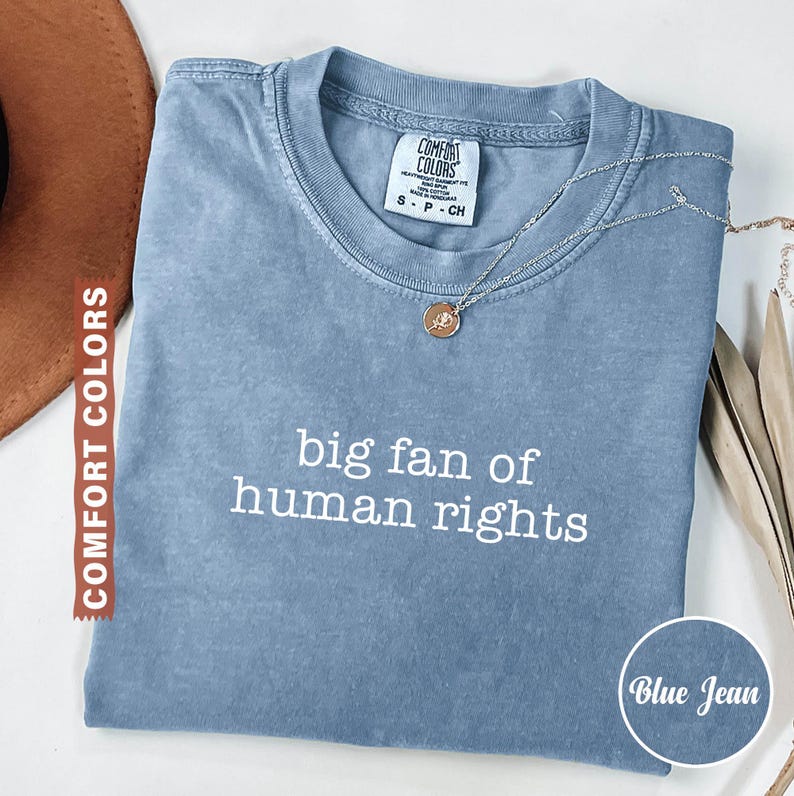 May include: A blue jean Comfort Colors t-shirt with the text "big fan of human rights" in white. A silver necklace with a pendant is on the shirt. The shirt is folded and displayed with a brown hat and dried leaves.