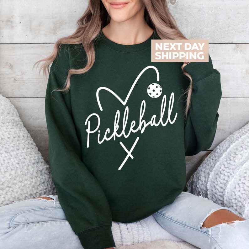 May include: A dark green sweatshirt with a white graphic of a heart containing a pickleball paddle and ball. The text "Pickleball" is written in a cursive font above the heart.