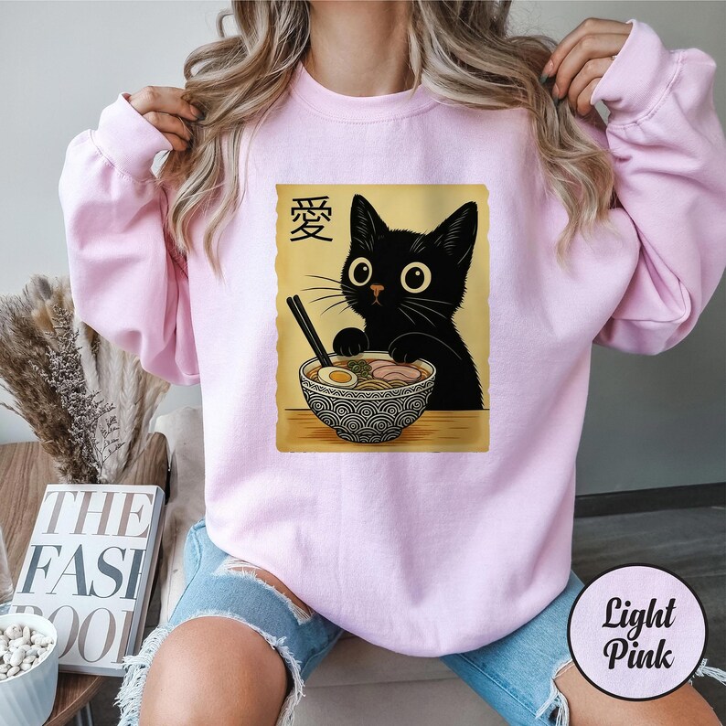 May include: Light pink sweatshirt featuring a black cat enjoying ramen. The cat is depicted with large eyes and is holding chopsticks. The ramen bowl has a decorative pattern. The Japanese character for "love" is above the cat.