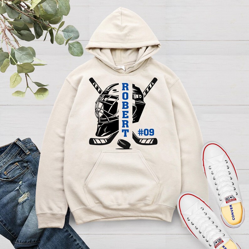 May include: A cream-colored hoodie with a hockey-themed design. The design features two hockey sticks crossed behind a hockey mask with the name "ROBERT" and the number "#09" printed on the hoodie.