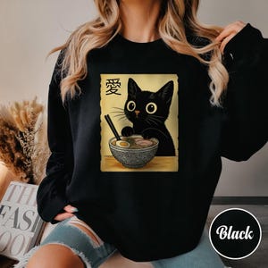 May include: Black sweatshirt featuring a cartoon cat with large eyes, holding chopsticks over a bowl of ramen. The design includes Japanese characters and is set on a tan background. The word "Black" is in a white circle.