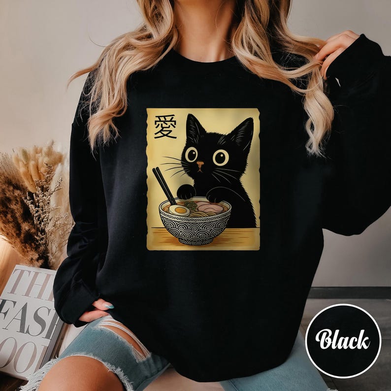 May include: Black sweatshirt featuring a cartoon cat with large eyes, holding chopsticks over a bowl of ramen. The design includes Japanese characters and is set on a tan background. The word "Black" is in a white circle.