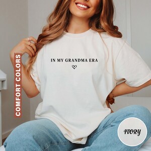 May include: Ivory-colored t-shirt with the text "IN MY GRANDMA ERA" and a heart symbol. The shirt is a Comfort Colors brand. The model is wearing blue jeans. The shirt is a crew neck.