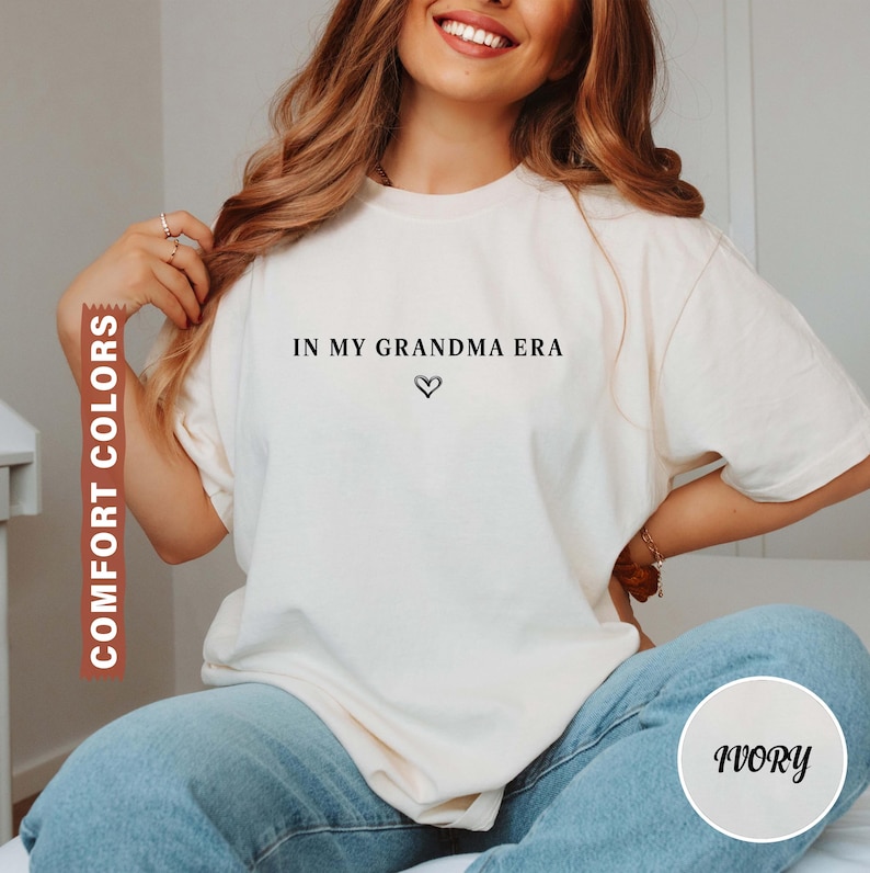 May include: Ivory-colored t-shirt with the text "IN MY GRANDMA ERA" and a heart symbol. The shirt is a Comfort Colors brand. The model is wearing blue jeans. The shirt is a crew neck.