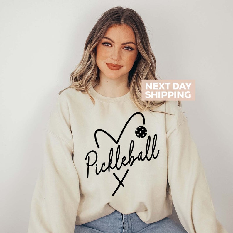 May include: A cream-coloured sweatshirt with a black graphic of a heart with a pickleball inside and the word "Pickleball" written in a cursive font.