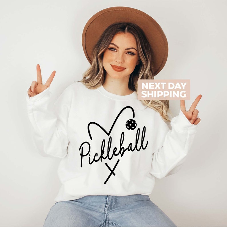May include: White sweatshirt featuring a black graphic design of a heart with a pickleball inside and the word "Pickleball" written in cursive below. The graphic also includes a pair of crossed pickleball paddles.