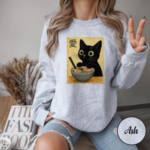 May include: Light gray sweatshirt featuring a graphic of a black cat with large eyes, eating ramen from a patterned bowl. The image includes Japanese text and is printed on a light gray sweatshirt.
