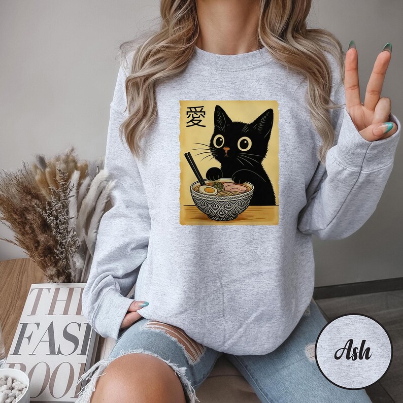 May include: Light gray sweatshirt featuring a graphic of a black cat with large eyes, eating ramen from a patterned bowl. The image includes Japanese text and is printed on a light gray sweatshirt.