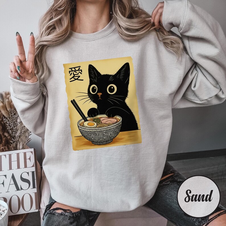 May include: A light gray sweatshirt featuring a graphic of a black cat eating ramen. The cat is depicted with large eyes and is holding chopsticks. The word "愛" is above the cat. The sweatshirt has long sleeves.
