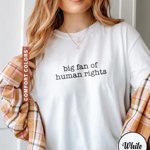 May include: White long-sleeve t-shirt with the text "big fan of human rights" in black. The model is wearing a plaid shirt over the t-shirt. The t-shirt is labeled "Comfort Colors" and "White".