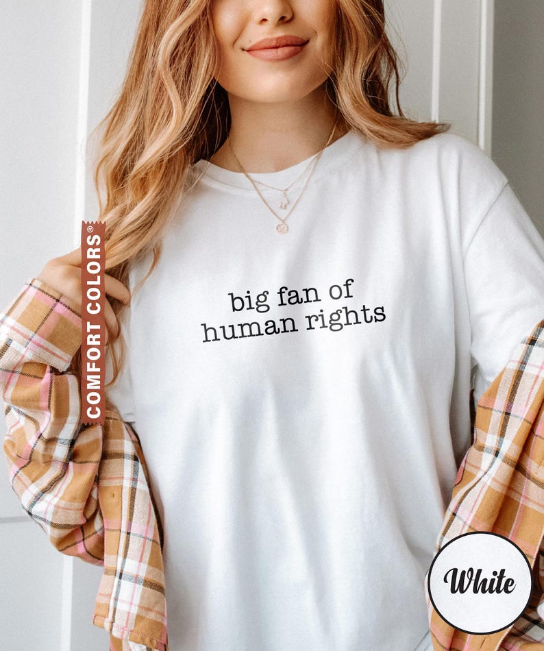 May include: White long-sleeve t-shirt with the text "big fan of human rights" in black. The model is wearing a plaid shirt over the t-shirt. The t-shirt is labeled "Comfort Colors" and "White".