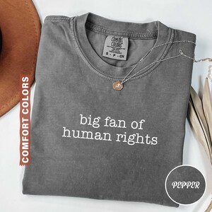May include: A gray Comfort Colors t-shirt with the text "big fan of human rights" in white. A silver necklace with a pendant rests on the shirt. The shirt is folded and displayed with a brown hat and dried plants.
