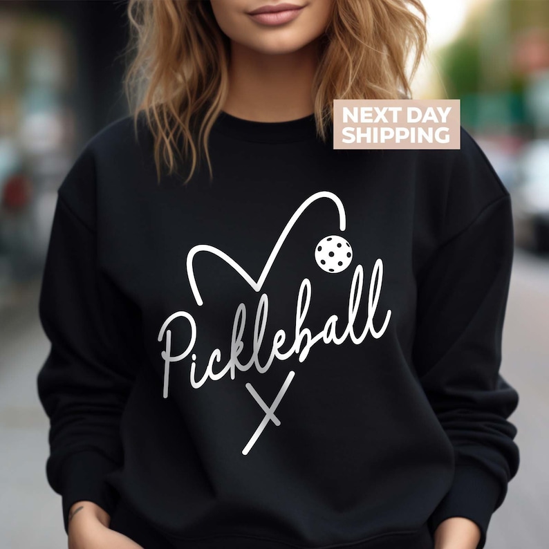 May include: Black sweatshirt with a white graphic design of a heart with a pickleball and paddles, and the text "Pickleball" in a cursive font.