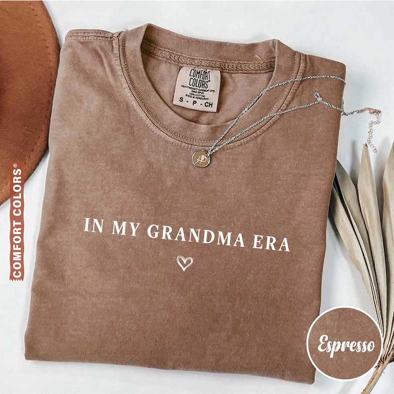 May include: Espresso-colored t-shirt with the text "IN MY GRANDMA ERA" and a heart symbol. The shirt has a crew neck and short sleeves. A silver necklace with a pendant is draped across the shirt.
