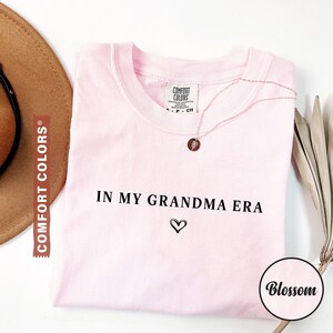 May include: A light pink Comfort Colors t-shirt with the phrase "IN MY GRANDMA ERA" and a heart symbol printed in black. A gold necklace with a pendant is draped on the shirt. A brown hat is visible on the left.