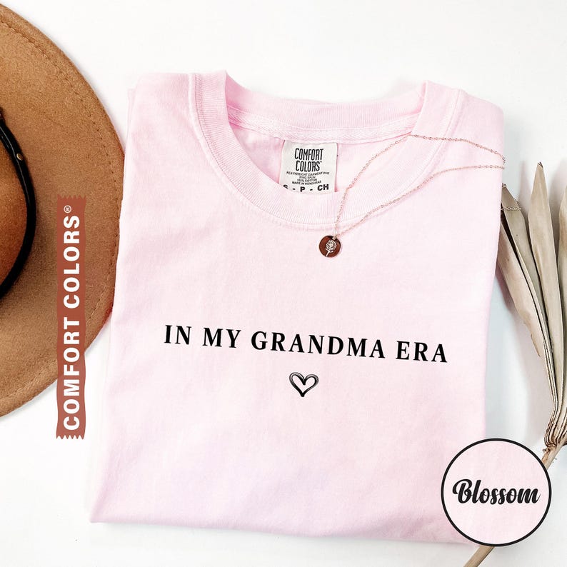 May include: A light pink Comfort Colors t-shirt with the phrase "IN MY GRANDMA ERA" and a heart symbol printed in black. A gold necklace with a pendant is draped on the shirt. A brown hat is visible on the left.