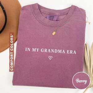 May include: Berry-colored t-shirt with the text "IN MY GRANDMA ERA" and a heart symbol. The shirt has a crew neck and a small Comfort Colors tag. A gold necklace with a rectangular pendant is draped across the shirt.
