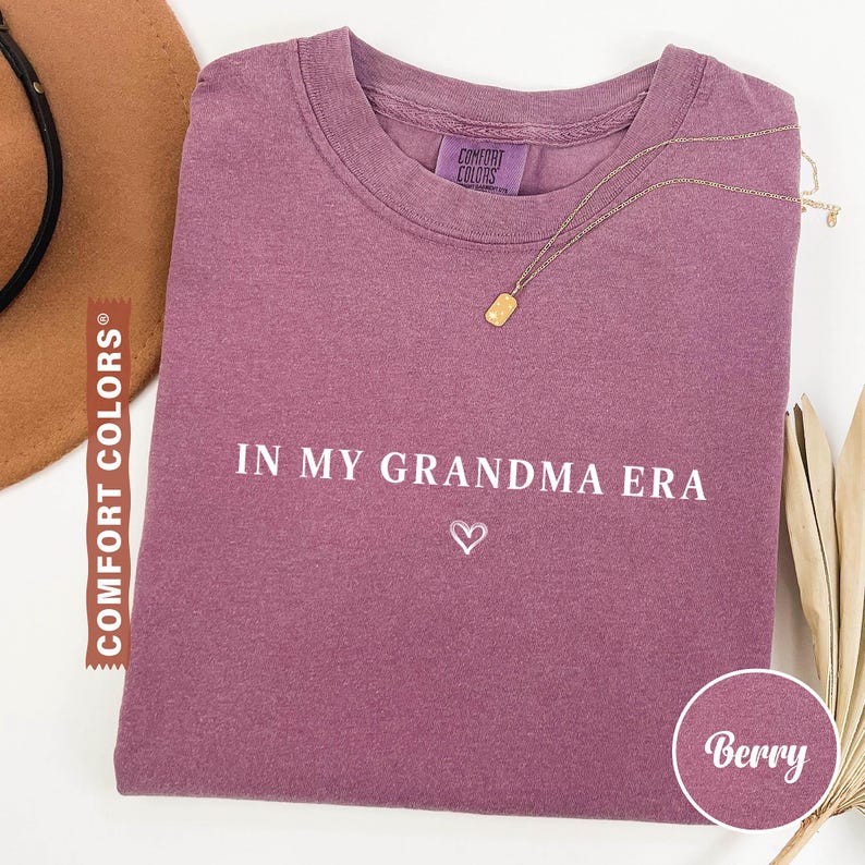 May include: Berry-colored t-shirt with the text "IN MY GRANDMA ERA" and a heart symbol. The shirt has a crew neck and a small Comfort Colors tag. A gold necklace with a rectangular pendant is draped across the shirt.