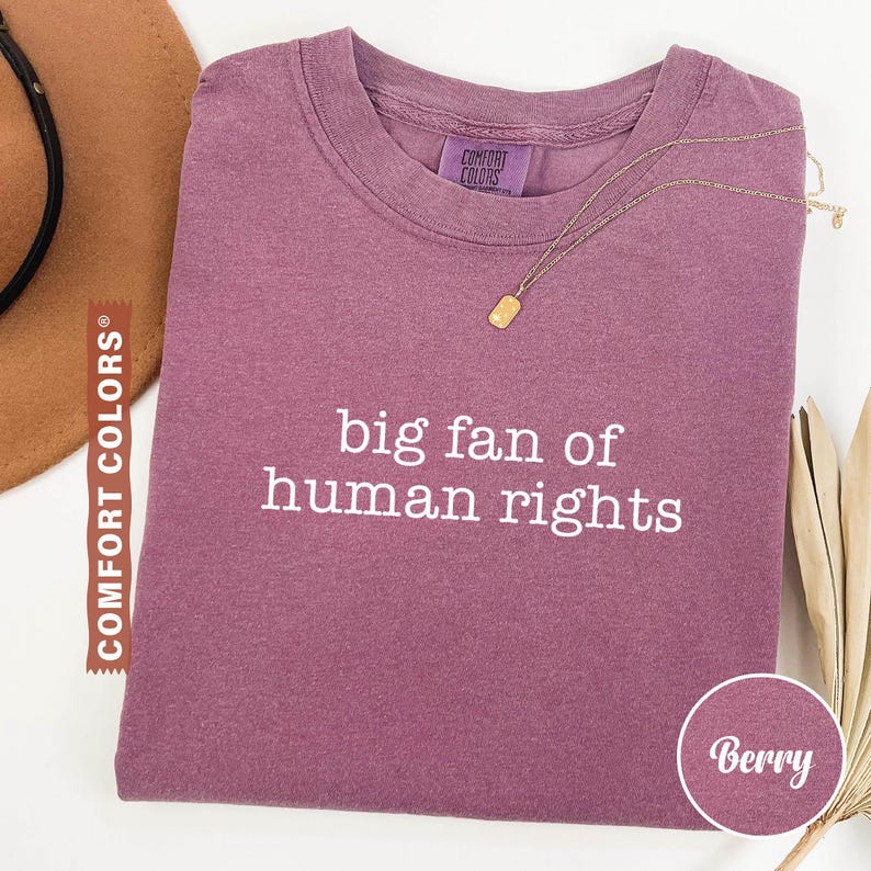 May include: Berry-colored t-shirt with the text "big fan of human rights" in white. The shirt has a crew neck and a small Comfort Colors tag. A gold necklace with a rectangular pendant rests on the shirt.