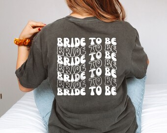 May include: A dark grey t-shirt with the text "BRIDE TO BE" repeated in a white, retro font.