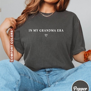 May include: Dark gray t-shirt with the text "IN MY GRANDMA ERA" and a small heart. The shirt is paired with blue jeans. The image also includes the words "COMFORT COLORS" and "Pepper".