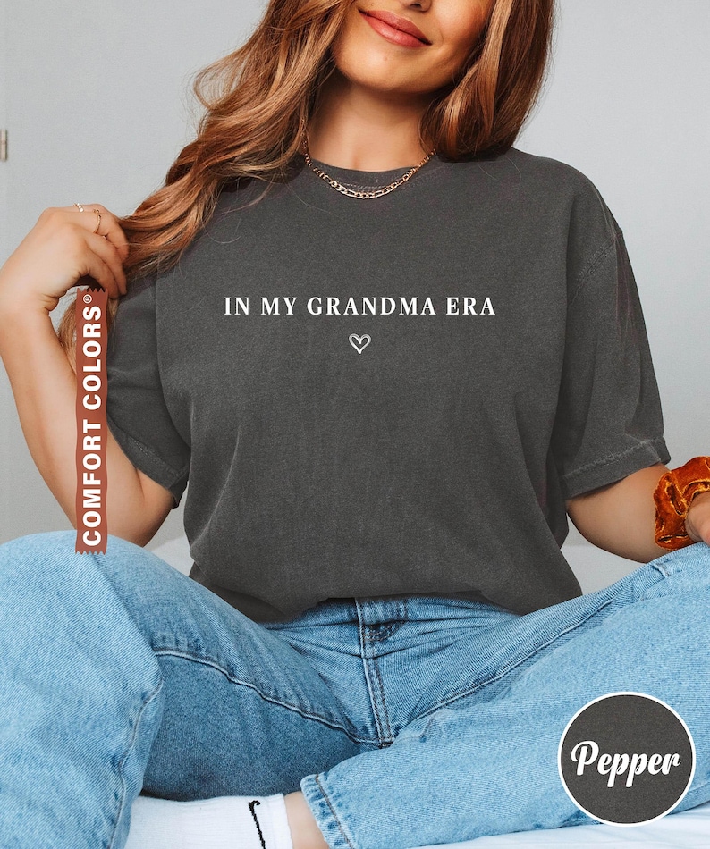 May include: Dark gray t-shirt with the text "IN MY GRANDMA ERA" and a small heart. The shirt is paired with blue jeans. The image also includes the words "COMFORT COLORS" and "Pepper".