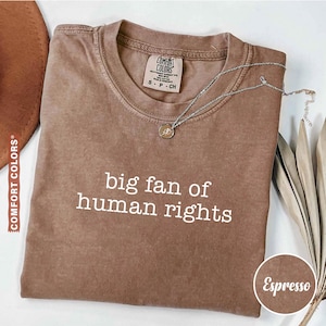 May include: Espresso-colored Comfort Colors t-shirt with the text "big fan of human rights" in white. A silver necklace with a pendant rests on the shirt. The shirt is folded and displayed with a brown hat and dried plant.