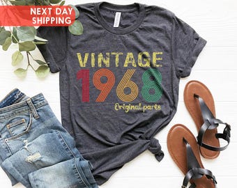 Puede incluir: Camiseta gris oscuro con el texto "VINTAGE 1968 Original parts" en amarillo, rojo, naranja y verde. La camiseta se combina con vaqueros azules y sandalias marrones con correas negras. Se ve vegetación en el fondo.