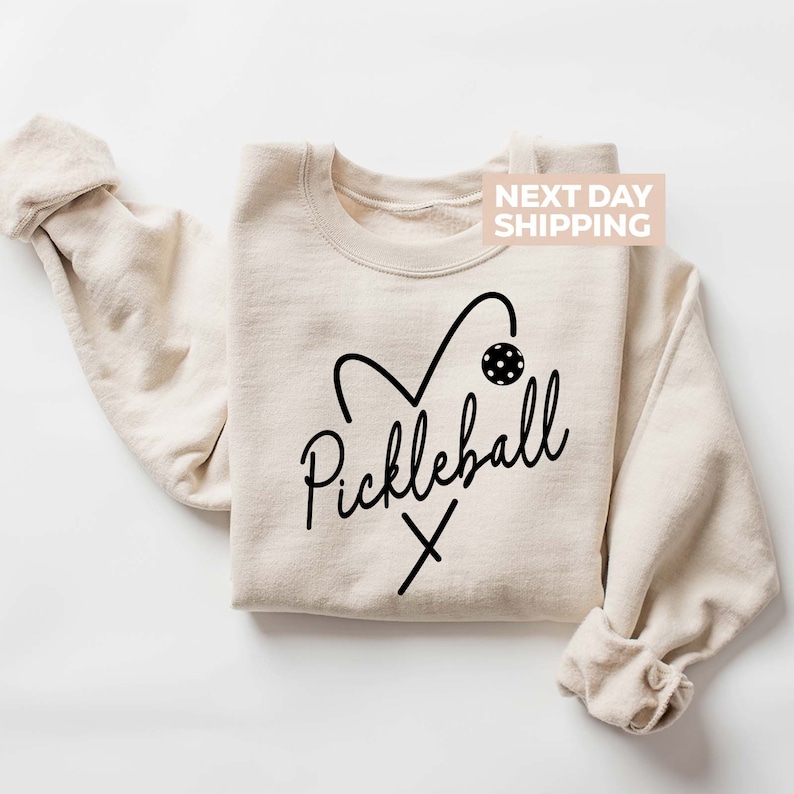 May include: A beige sweatshirt with a black heart design that includes a pickleball and paddles. The text 'Pickleball' is written in a cursive font below the heart. The text 'NEXT DAY SHIPPING' is in a white rectangle in the upper right corner of the image.