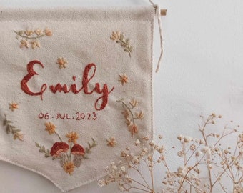 May include: A beige canvas banner with embroidered flowers, mushrooms, and the name "Emily" in red cursive lettering. The date "06. JUL. 2023" is also embroidered below the name.