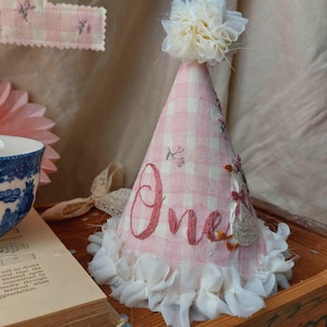 Peut inclure: Un chapeau de fête rose et blanc à carreaux vichy avec le mot "One" brodé sur le devant. Le chapeau est orné de volants blancs et surmonté d'une fleur blanche. Une bannière florale décorative est visible en arrière-plan.