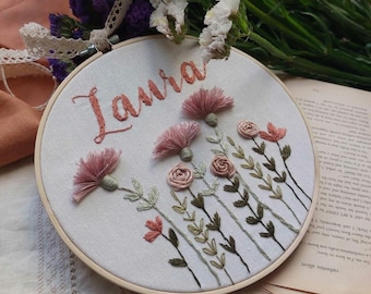 May include: Embroidered hoop art with the name "Laura" stitched in cursive script. The hoop features a floral design with pink flowers and green leaves.