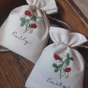 Könnte beinhalten: Zwei cremefarbene Stoffbeutel mit gestickten roten Erdbeeren, grünen Blättern und weißen Blüten. Auf jedem Beutel ist der Name "Emily" in brauner Schrift gestickt. Die Beutel sind mit Bindfäden gebunden, geeignet für kleine Geschenke oder Säckchen.