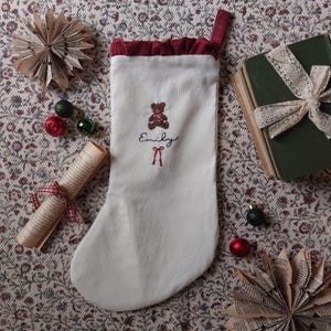 May include: A white fabric Christmas stocking featuring a dark red ruffled cuff and a hanging loop. The stocking is embroidered with a brown teddy bear, the name Emily in black script, and a small red bow detail below the name.