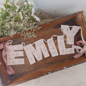 Może przedstawiać: Baner z napisem "EMILY" w różowych literach z tkaniny w kratkę z kwiatowymi akcentami. Litery połączone są białym sznurkiem i ozdobione różowymi wstążkami. Baner spoczywa w rustykalnej drewnianej tacy, z białymi kwiatami w tle.