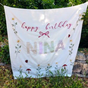 Pode incluir: Uma faixa de tecido branco com o texto "happy birthday" em letra cursiva vermelha acima de um laço vermelho. O nome "NINA" é exibido em letras grandes e estampadas. O design inclui margaridas, joaninhas e cogumelos bordados.