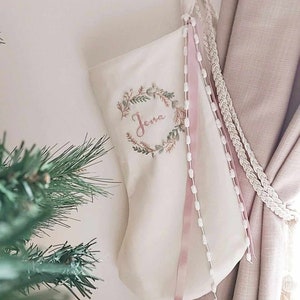 May include: Cream Christmas stocking embroidered with the name "Jena" within a wreath. Pink ribbons and white beaded accents adorn the stocking, which hangs near a curtain.