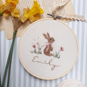 Può includere: Arte di ricamo su un cerchio che rappresenta un coniglio marrone con un nastro rosa, circondato da fogliame verde e fiori rosa. Il nome "Emily" è ricamato sotto il coniglio.
