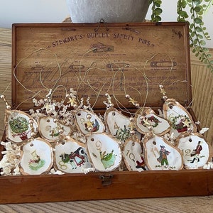 May include: A wooden box with a hinged lid, containing 12 oyster shell ornaments with gold wire hangers. Each ornament is painted with a different Christmas scene and numbered 1 through 12. The box is labeled "Stewart's Duplex Safety Pins".